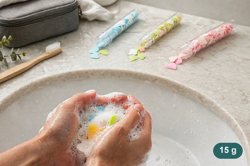 Hands lathering a soap sheet at a sink, with three bottles and a wash bag nearby